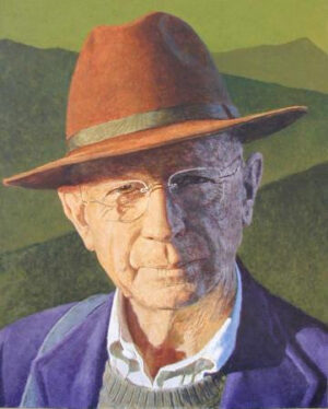 Portrait of an elderly man wearing a brown fedora, glasses, a collared shirt, and a blazer against a hilly landscape.