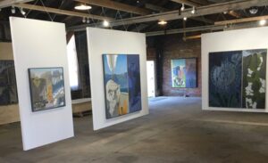 Paintings displayed on white partitions in an industrial gallery space with exposed brick walls.