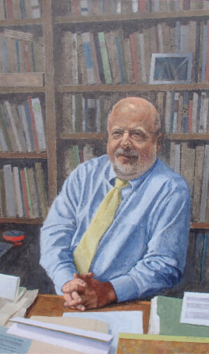 A portrait of a bald, bearded man in a blue shirt and yellow tie sitting at a desk in front of a bookshelf.